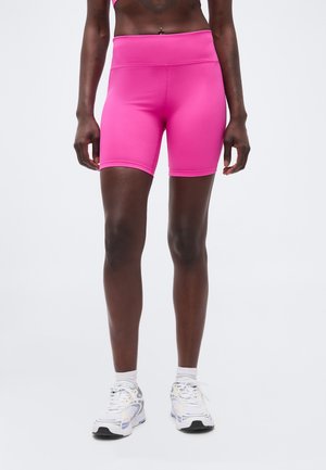 Kobieta ubrana w jasnoróżowe, wysokie krótkie spodenki rowerowe, białe skarpetki i białe adidasy, stojąca na tle jednolitego jasnego tła.