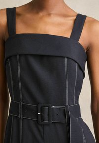 Zwarte jurk met brede bandjes en een vierkante halslijn, voorzien van witte stiksels en een ceintuur in de taille. Glad materiaal met gestructureerde designelementen.