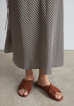 Personne portant des sandales marron en daim à brides croisées et une jupe midi à carreaux noirs et blancs, debout sur un sol en béton.