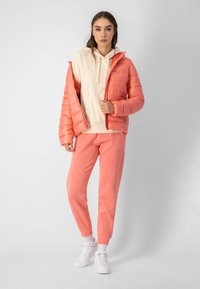 Gepolsterte korallenfarbene Jacke mit Kapuze, über einem cremfarbenen Hoodie getragen, kombiniert mit korallenfarbenen Sweatpants und weißen Sneakers. Glatte Textur, lässiges Design.