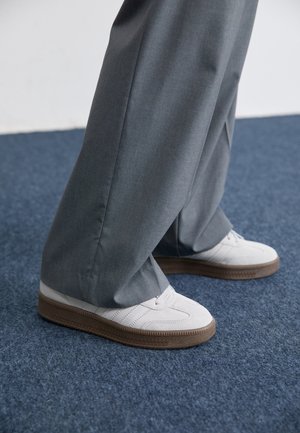 Scarpe da ginnastica bianche con suole marroni abbinate a pantaloni grigi larghi, in piedi su un tappeto blu testurizzato.