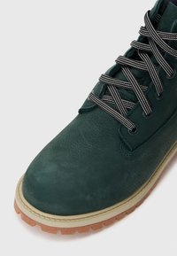 Stivale verde in pelle con superficie testurizzata, lacci a strisce bianche e nere e suola beige, dotato di un battistrada robusto.