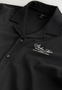 Chemise noire boutonnée avec un large col et texte brodé blanc « Studios Italia ESTD. MCMXL » sur la poitrine, fabriquée par la marque NEXT.