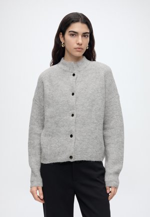 Femme aux cheveux foncés portant des boucles d’oreilles créoles dorées, un pull gris clair boutonné et un pantalon noir sur un fond blanc uni.