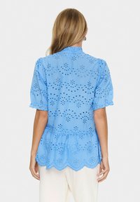 Blusa azzurro chiaro con motivi ricamati a giorno, maniche corte a sbuffo, colletto arricciato e orlo ondulato. Tessuto leggero.