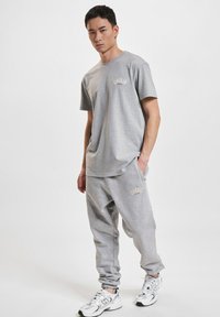 Szary bawełniany t-shirt i dopasowane dresy z ściągaczami. Logo "UNFAIR" w złotej haftowanej wersji. Model nosi białe buty sportowe.