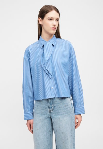 Γυναίκα που φοράει μπλε πουκάμισο με ρίγες και μακριά μανίκια με λεπτομέρεια φουλαριού στο λαιμό και ανοιχτόχρωμο τζιν ψηλόμεσο διακοσμημένο με μικρές τρουκς.