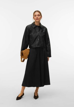 Femme portant une veste en cuir noire courte, une longue jupe noire, des talons foncés, tenant un sac brun plié devant un fond blanc uni.