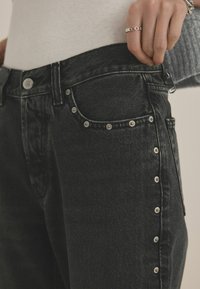 Mustmustest denim püksid, mis on kõrge vöökohaga, hõbedase nupuga sulgemine ja dekoratiivsed metallkliibid külgõmblusel. Nähtav tekstuuriga kangas.