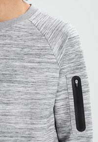 Sudadera gris texturizada con un patrón de rayas horizontales. Cuenta con un bolsillo con cremallera negro en la manga izquierda y un cuello redondo.