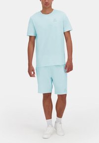 Camiseta de algodón azul claro con mangas cortas y cuello redondo, combinada con pantalones cortos a juego que presentan un logo. Zapatillas blancas completan el conjunto.