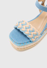 Blue denim wedge sandal with a braided cream and blue strap. Features a textured espadrille sole with a woven design and a smooth inner lining.