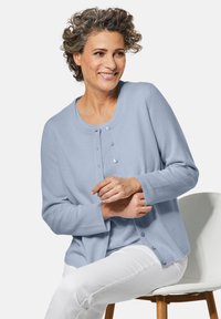 Hellblauer Cardigan aus weichem Material mit Rundhals, durchgehender Knopfleiste und gerippten Bündchen, kombiniert mit weißen Hosen.