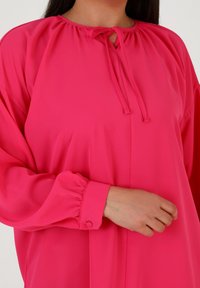 Blusa rosa brillante con scollatura arricciata e chiusura con cravatta. Presenta maniche lunghe con polsini elastici e un tessuto morbido e leggero.