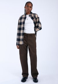 Ruitjes flanellen shirt in marineblauw en beige over een wit tanktop, gecombineerd met bruine wijde broek en zwarte schoenen. Effen achtergrond.