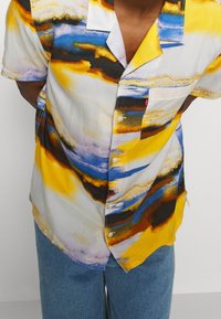 Kortärmad skjorta med ett mångfärgat abstrakt mönster med gula, blå och svarta accenter. Lättviktsmaterial med en fickdetalj.