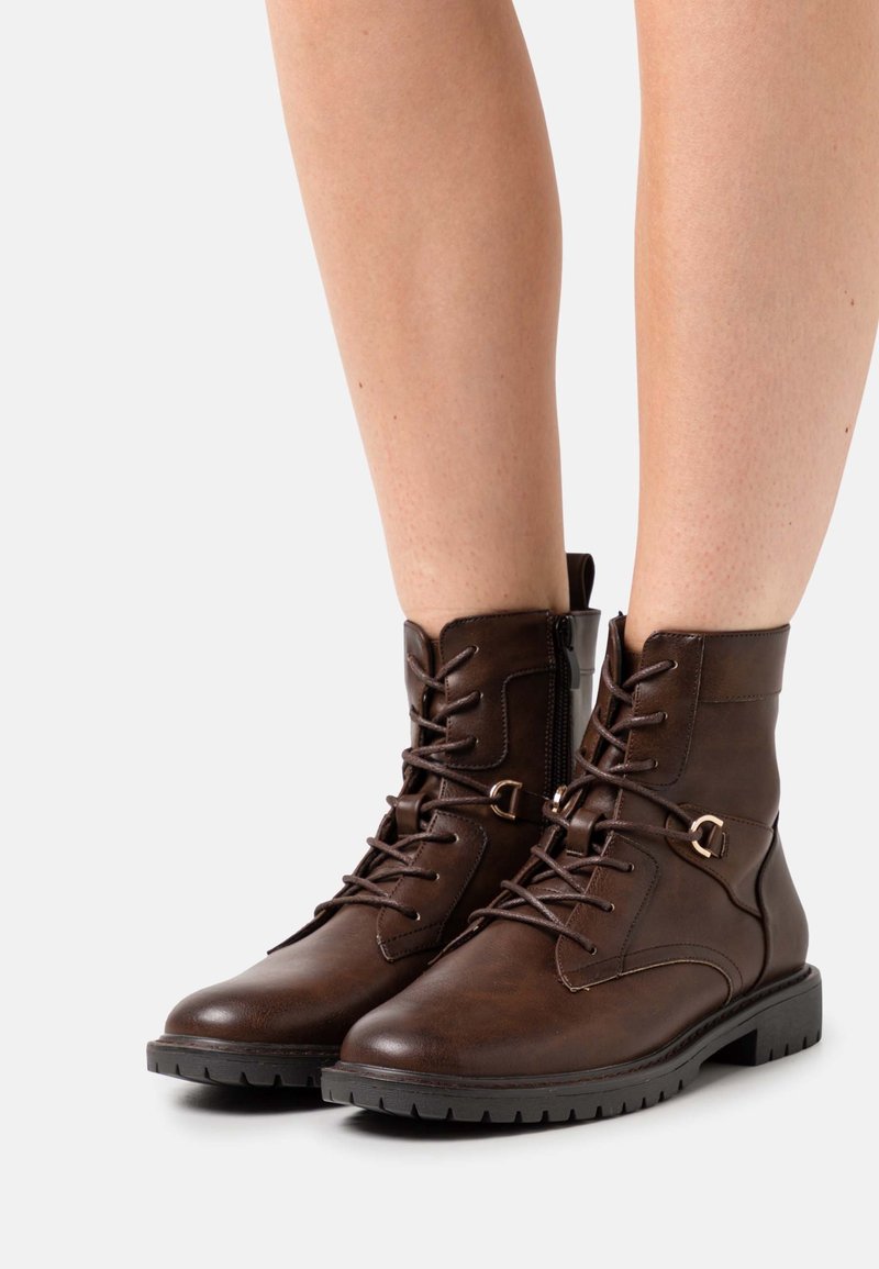 Bottes basses en cuir marron à lacets avec fermeture éclair latérale, portées sur des jambes nues, dotées de semelles en caoutchouc noir et d'œillets métalliques décoratifs.