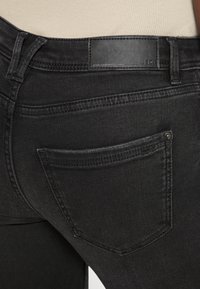 Jeans de mezclilla negra con una textura suave, que cuentan con un bolsillo trasero y una etiqueta de cuero en la cintura. Vista en primer plano de los detalles de costura.