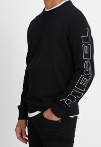 Sudadera negra de manga larga con puños acanalados y cuello redondo. Presenta un prominente logo blanco "DIESEL" en la manga izquierda.