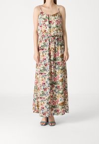 Femme portant une robe maxi fleurie sans manches avec fines bretelles et taille élastique, associée à des talons ouverts bleus.