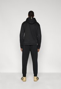 Μαύρο φούτερ με υφασμένο κουκούλα και ριμπ τελείωμα, συνδυασμένο με αντίστοιχο παντελόνι φόρμας· άτομο που φοράει ανοιχτόχρωμα παπούτσια με σχέδια.