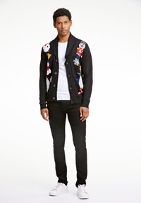 Giovane uomo che indossa un cardigan di Natale nero con motivi festivi, una maglietta bianca, pantaloni neri e sneakers bianche, in piedi su uno sfondo bianco.