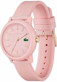Montre en silicone rose avec un bracelet texturé, des accents dorés et un cadran rond. Présente un logo Lacoste et un cadran minimaliste avec des index horaires dorés.