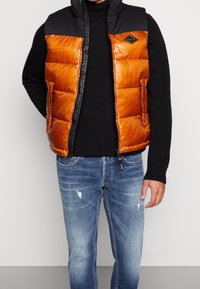 Vadderad väst med en orange reflekterande utsida och en svart ovandel. Har en frontdragkedja och sidofickor, bärs över en svart tröja.
