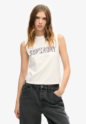 Biały, bezrękawnikowy crop top z nadrukiem "Superdry" w teksturowanym wzorze, w połączeniu z ciemnoszarymi spodniami z wysokim stanem, posiadającymi przednie kieszenie.
