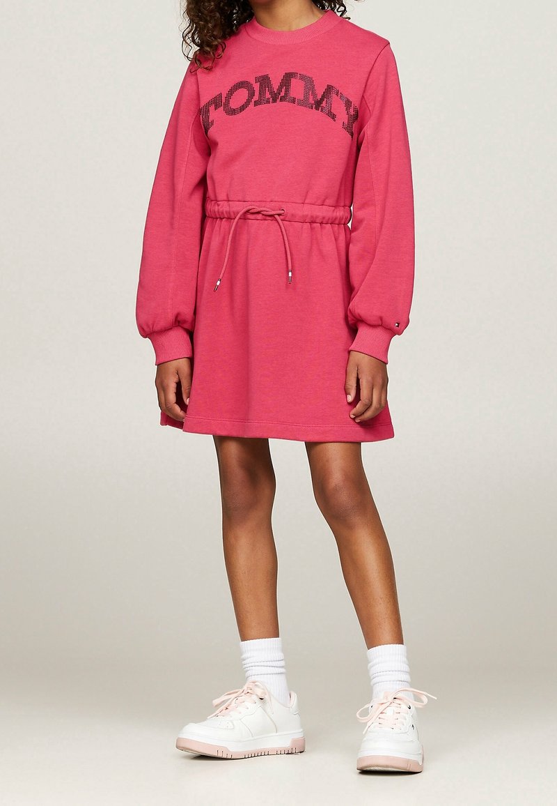 Enfant portant une robe rose à manches longues avec le texte « TOMMY », taille à cordon, chaussettes blanches et baskets blanches, debout sur un fond neutre.