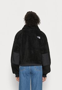 Giacca in pile nera con una texture morbida, design oversize, colletto alto e dettagli in nylon nero sulle maniche. Presenta un logo sul retro.