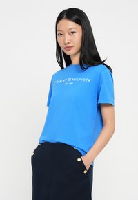 Młoda kobieta z prostymi czarnymi włosami nosi niebieską koszulkę Tommy Hilfiger i ciemną spódnicę z złotymi guzikami, z rękami w kieszeniach, na neutralnym tle.