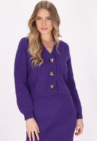 Violetti neulecardigan, jossa on V-pääntie, kolme suurta nappia ja teksturoitu sahalaita-kuvio. Muotoiltu malli pitkillä hihoilla.