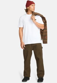 Witte T-shirt, bruine cargo broek, rode gebreide beanie, houdt een gebloemde jas vast. Zwarte sneakers, relaxte pasvorm, zichtbare zakdetails op de broek.