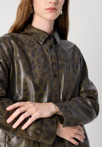 Giacca camicia in pelle verde oliva con stampa leopardata nera, chiusura con bottoni, colletto e maniche lunghe con polsini abbottonati.