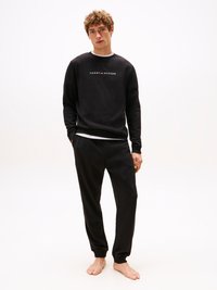 Melnā džemperis ar balto "TOMMY HILFIGER" logotipu, kombinēts ar melniem sporta biksēm. Mīksta auduma, ribota piegriezuma piedurknes un apakšmalas. Modelis stāv basām kājām.