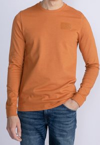 Långärmad orange t-shirt i bomull, med en liten rektangulär logotyp på bröstet och rundad halsringning.