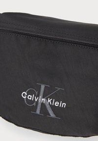 Čierna nylonová pásová taška s uzatváraním na zips, s embosovaným logom "CK" a textom "Calvin Klein" v bielej farbe, s hladkou textúrou a zaobleným tvarom.