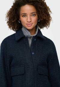 Mornarice plavi teksturirani kaput s ovratnikom, dva prednja džepa i kopčama na dugmad. Nositi preko sive majice s visokim ovratom.