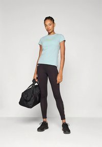 Světlomodré přiléhavé tričko, černé legíny a černé tenisky. Modelka drží černou sportovní tašku s popruhem. Legíny mají bílé textové detaily.