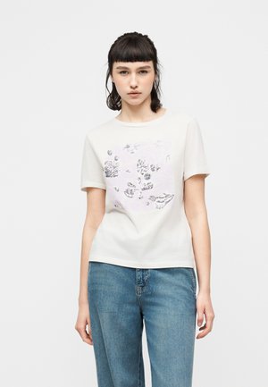 Junge Frau mit dunklen Haaren trägt ein weißes Kurzarmshirt mit silbernem Blumenmuster und blaue Jeans, steht vor einem schlichten weißen Hintergrund.