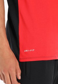 Camiseta deportiva Dri-FIT en rojo y negro con mangas cortas, que cuenta con una textura suave y un logo en la parte inferior derecha.