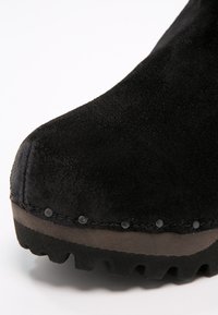 Bottine en suède noir avec un bout arrondi, une surface texturée et une semelle en caoutchouc robuste. Présente de petites rivets décoratifs le long de la couture.