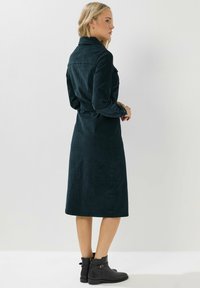 Abito lungo verde scuro in suede con colletto a bottoni e cintura in vita. La modella indossa stivaletti neri alla caviglia e si trova di spalle rispetto alla fotocamera.