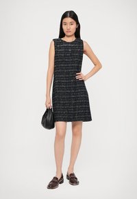Vestido sem mangas azul-marinho com um tecido texturizado, apresentando detalhes dourados. A modelo segura uma bolsa preta e usa sapatos escuros e brilhantes com um salto baixo.