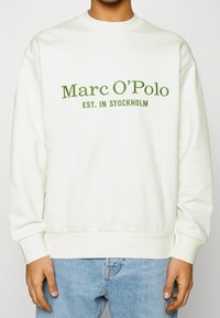 Sweat-shirt couleur crème avec texte brodé en vert indiquant "Marc O'Polo EST. IN STOCKHOLM", avec un col rond et des poignets côtelés.