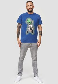 Blauwe katoenen T-shirt met een grote Goofy-afbeelding in zwart en groen. Gekocht in combinatie met lichtgrijze jeans en witte sneakers. Korte mouwen, ronde hals.