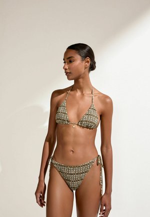 Mujer con cabello oscuro recogido en un moño bajo, vestida con un bikini de cordones con estampado verde y blanco, de pie frente a un fondo beige liso.