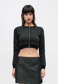 Cardigan negru scurt din tricot cu model de logo argintiu, manșete striate și închidere frontală cu fermoar, asortat cu o fustă mini din denim închis la culoare.