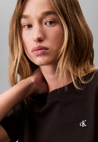 Junge Frau mit hellbraunen Haaren, die ein schwarzes Calvin Klein T-Shirt trägt und eine Hand am Hals anlehnt, neutraler Hintergrund.